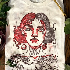 May include: White t-shirt with a red and black linocut print of a woman's face with a butterfly, snake, and flowers. The woman's hair is half red and half black.