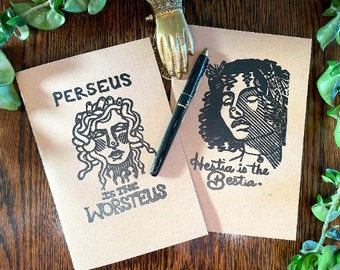 Puede incluir: Dos cuadernos de papel marrón con ilustraciones de tinta negra. El cuaderno de la izquierda tiene una ilustración en blanco y negro de una mujer con serpientes en el pelo y el texto "Perseus is the Worsteus". El cuaderno de la derecha tiene una ilustración en blanco y negro de la cara de una mujer y el texto "Hestia is the Bestia."