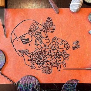 May include: A black and white linocut print on orange paper featuring a skull with a butterfly and flowers. The skull has a heart-shaped eye.