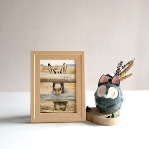 Op de afbeelding: Een houten fotolijst met een foto van een ezel die door een houten hek kijkt. De ezel is bruin en wit met grote oren. De lijst staat op een wit oppervlak naast een grijze keramische vaas met een roze en wit gezicht.