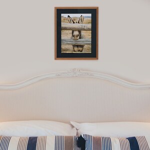 Op de afbeelding: Een ingelijste foto van een ezel die door een houten hek kijkt. De ezel is grijs en wit met grote oren. De foto hangt aan een witte muur boven een wit hoofdbord met een wit en blauw gestreept kussen.