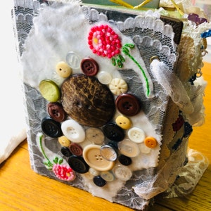 May include: A handmade journal with a grey cover decorated with a variety of buttons, a coconut shell, and a pink embroidered flower. The journal is wrapped in white lace and fabric.