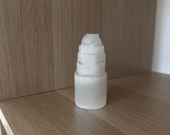 Könnte beinhalten: Eine weiße Selenit-Turmskulptur mit einem geschichteten Design. Der Kristall ist an der Basis zylindrisch, mit gestapelten, progressiv kleiner werdenden Ebenen nach oben. Die Skulptur wird auf einem Holzregal ausgestellt.