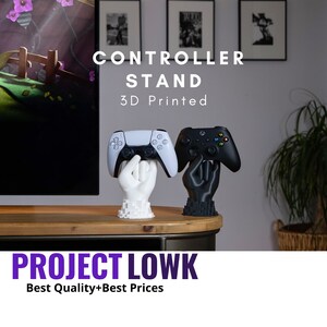May include: A 3D printed controller stand with a white hand holding a white PlayStation 5 controller and a black hand holding a black Xbox controller. The stand is on a wooden table. The text "CONTROLLER STAND 3D Printed" is above the stand. The text "PROJECT LOWK Best Quality+Best Prices" is below the stand.