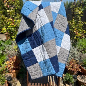 May include: A handmade quilt with a patchwork design in shades of blue, gray, and white. The quilt is draped over a wooden chair in a garden setting. The quilt features a variety of geometric shapes and patterns, with a dark blue border.