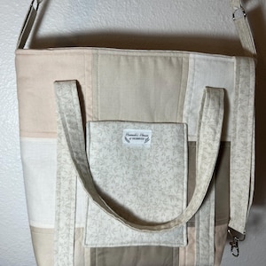 May include: A beige and cream-colored tote bag with a patchwork design. The bag features a front pocket with a floral pattern and the text "Hannah's House of Hobbies". It has a shoulder strap and two handles.