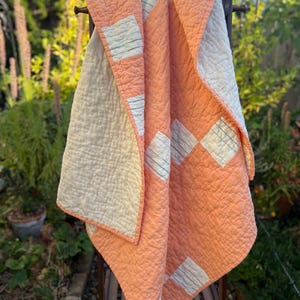 May include: A quilted blanket featuring a geometric pattern of peach-colored diamonds and off-white diamond shapes. The off-white sections are detailed with thin, dark horizontal lines. The fabric has a visible, crinkled texture throughout the quilt.