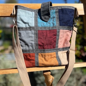 May include: A patchwork shoulder bag with a beige canvas strap. The bag features a variety of colorful fabric squares in shades of blue, gray, red, and tan, stitched together with gray thread. A dark gray flap secures the top.