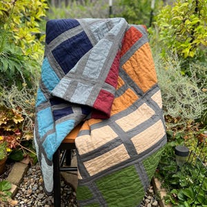 May include: A patchwork quilt featuring a grid of rectangular fabric panels in navy blue, light blue, red, orange, beige, and olive green, all separated by dark gray sashing. The textile has a visible quilted texture and folded edges.