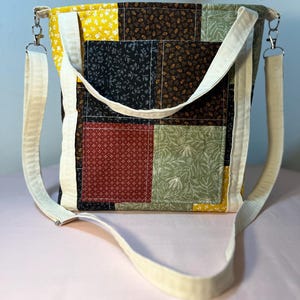 以下が含まれることがあります： クリーム色のショルダーストラップとハンドルが付いたパッチワークトートバッグ。黄色、黒、緑、赤の柄の生地の正方形が特徴です。バッグは長方形で、物を運ぶのに適しています。