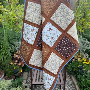 May include: A patchwork quilt featuring alternating square blocks of brown fabric with small white floral patterns, tan fabric with thin brown stripes, and light blue fabric printed with small orange dragons and yellow castles, all bordered by light blue trim.