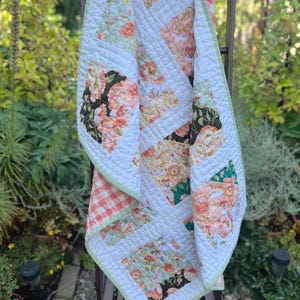 May include: A patchwork quilt featuring a mix of floral patterns in shades of peach, green, and black, interspersed with solid white fabric blocks and a light orange gingham border. The quilt has a quilted texture and a light green fabric binding.