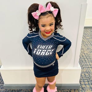 May include: A navy blue cheerleading uniform featuring the text CHEER FORCE in white letters on the chest, accented with silver rhinestones. The outfit includes a long-sleeved top, a matching skirt, pink hair bows, and white trainers with pink ruffled socks.