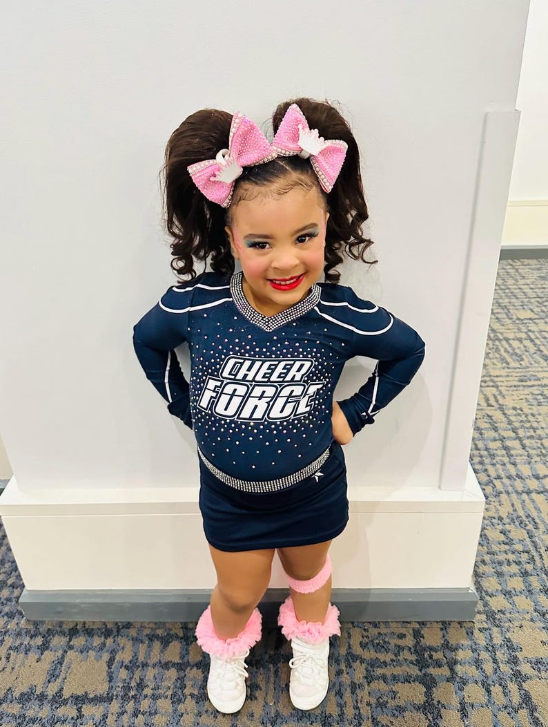 May include: A navy blue cheerleading uniform featuring the text CHEER FORCE in white letters on the chest, accented with silver rhinestones. The outfit includes a long-sleeved top, a matching skirt, pink hair bows, and white trainers with pink ruffled socks.