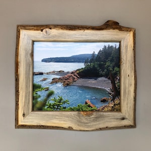 May include: A rustic wooden picture frame with a natural wood finish. The frame features a landscape photograph of a rocky coastline with a small cove of blue water.
