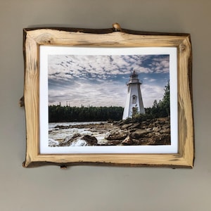 May include: A framed print of a white lighthouse on a rocky shoreline under a cloudy sky. The frame is made of natural-edge wood. The lighthouse has a red top and is surrounded by green trees.