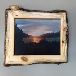 May include: A rectangular picture frame crafted from light-coloured wood with a natural live edge, featuring dark bark along the outer perimeter and visible wood knots. The frame holds a landscape photograph of a sunset over a lake and mountains.