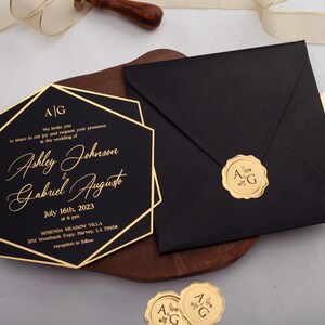 Puede incluir: Invitación de boda negra con borde geométrico dorado y texto. La invitación incluye los nombres Ashley Johnson y Gabriel Augusto, y la fecha 16 de julio de 2023. También hay un sobre negro con un sello dorado.