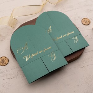 Può includere: Due partecipazioni di nozze verde acqua con scritta dorata, che recitano "We found our forever". Gli inviti sono su un vassoio di legno, con sigilli di cera dorati e un nastro. È visibile anche un timbro di legno.