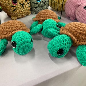 May include: Handmade crochet turtles with brown shells and green bodies, legs, and heads. The turtles have large black eyes and are displayed on a light-colored surface. Other plush animals are visible in the background.