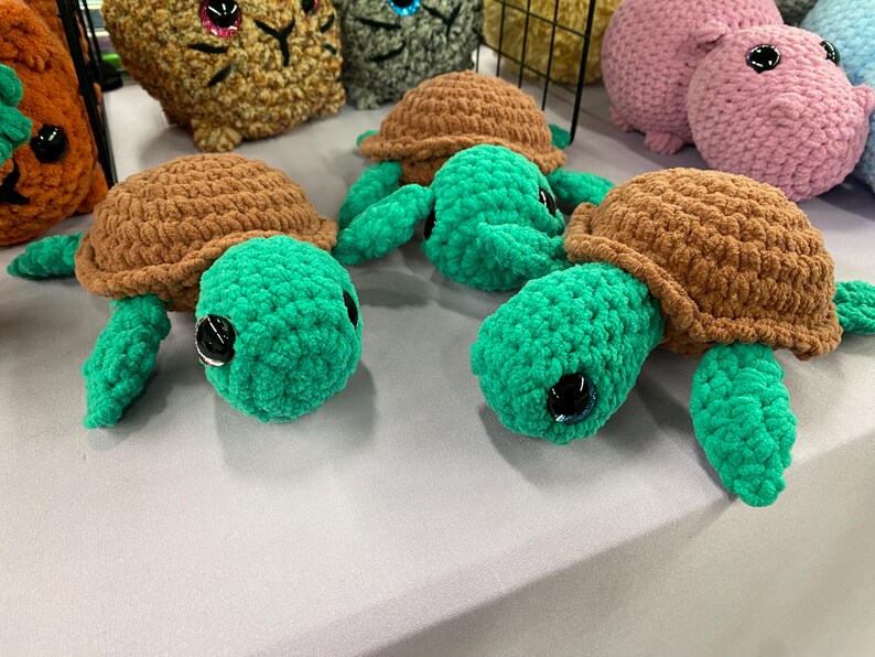 May include: Handmade crochet turtles with brown shells and green bodies, legs, and heads. The turtles have large black eyes and are displayed on a light-colored surface. Other plush animals are visible in the background.