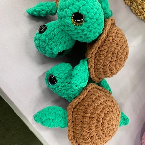 May include: Two handmade crochet sea turtles. The turtles have green heads, flippers, and limbs, with brown shells. The eyes are black with gold glitter accents. The turtles are made of soft, textured yarn.