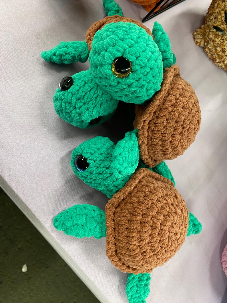 May include: Two handmade crochet sea turtles. The turtles have green heads, flippers, and limbs, with brown shells. The eyes are black with gold glitter accents. The turtles are made of soft, textured yarn.
