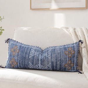 Puede incluir: Un cojín lumbar rectangular de tela azul intenso con un patrón de rayas verticales blancas y un intrincado bordado geométrico en tonos marrón y blanco. El cojín está rematado con pequeñas borlas de color azul oscuro en cada esquina.