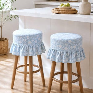 May include: Two round bar stools with light wood legs and circular footrests. The seats are upholstered in a light blue fabric featuring a white floral pattern, finished with a gathered fabric ruffle skirt around the edge of the seat cushion.