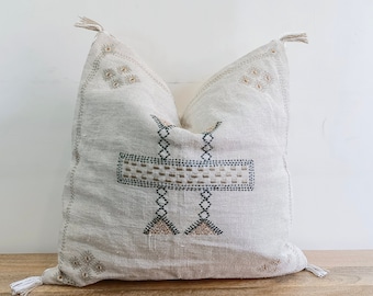 May include: A square beige throw pillow featuring intricate dark gray and tan embroidered geometric patterns on the front. The pillow has small tassels at each of the four corners and a textured fabric surface with visible woven threads throughout.