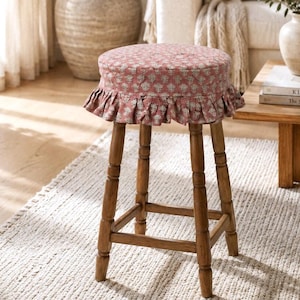 May include: A round wooden stool featuring a padded seat upholstered in a dusty rose fabric with a repeating white floral pattern and a gathered ruffled skirt. The stool has four turned wooden legs connected by a square support frame.