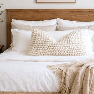 May include: A rectangular lumbar pillow with a cream-colored textured surface made of small, densely packed spherical pom-poms. The pillow is centered on a bed with white linens, beige pillows, and a beige fringed throw blanket.