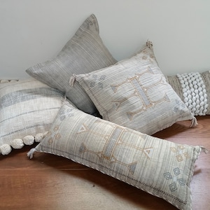 May include: Four decorative throw pillows with a neutral color palette. The pillows are made of a soft, textured fabric and feature intricate patterns in shades of gray, white, and beige. The pillows are arranged on a wooden floor.