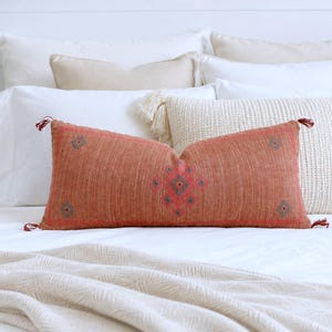 May include: A rectangular lumbar pillow in a burnt orange hue featuring a central diamond-shaped embroidery pattern in red and blue. The pillow has small tassels at each corner and is positioned in front of several white and beige decorative pillows.
