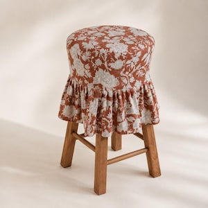 Op de afbeelding: Een ronde houten kruk met een gestoffeerde zitting bekleed met een roestkleurige stof met een wit bloemenpatroon. De zitting is voorzien van een geplooide, gerimpelde rok die over de vier rechte houten poten en verbindingsbalken valt.