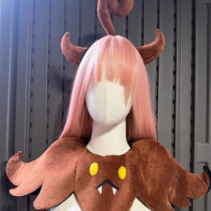 May include: Brown bat-shaped costume collar with yellow eyes and white fangs. The collar has a brown furry texture and is worn on a mannequin with pink hair and brown horns.