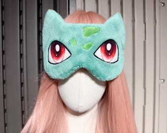 May include: A plush sleep mask in the shape of a Bulbasaur, featuring a light teal color with green accents and large red eyes. The mask is designed to cover the eyes and is displayed on a mannequin head with pink hair.