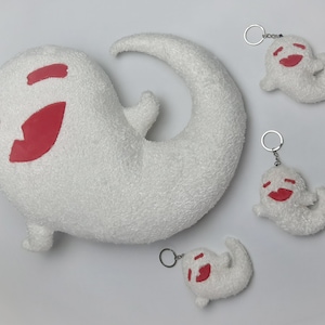 Puede incluir: Cuatro llaveros de peluche con forma de fantasma en blanco con caras sonrientes rojas. El fantasma más grande es una almohada con una cola curva. Los otros tres fantasmas son más pequeños y están unidos a llaveros.