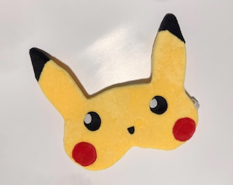 May include: A yellow plush Pikachu character with black-tipped ears, black eyes, and red cheek circles. The plush toy is a popular character from the Pokémon franchise.