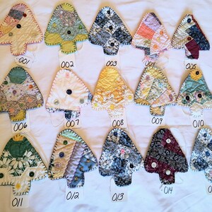 May include: Fifteen fabric ornaments shaped like Christmas trees, each featuring unique patchwork patterns, varied color palettes, and decorative buttons. Each ornament is labeled with a small white tag numbered from 001 to 015.