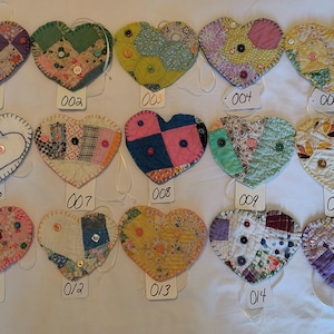 May include: Fifteen heart-shaped fabric ornaments arranged in three rows. Each heart features patchwork patterns, various floral or geometric prints, and small buttons sewn onto the surface. Each ornament has a white tag labeled 001 through 015.