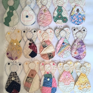 May include: A collection of fifteen quilted fabric angel ornaments, each labeled with a number from 001 to 015. These ornaments feature various patchwork patterns, colors, and textures, with small buttons attached to the center of each angel body.