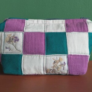 Op de afbeelding: Een gewatteerde make-uptas met een patchworkontwerp. De tas heeft vierkanten van teal, paarse en witte stof. Sommige witte vierkanten hebben bloemenborduurwerk. De tas heeft een ritssluiting en staat op een houten oppervlak.