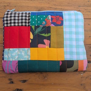 Op de afbeelding: Een rechthoekig tasje met ritssluiting, gemaakt van een patchwork van verschillende stoffen vierkanten en rechthoeken. Het ontwerp bevat een mix van patronen, waaronder zwart-wit gingham, bloemenprints, effen rode, gele, groene en lichtblauwe vlakken.