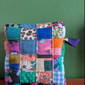 May include: A patchwork square pouch with a variety of colorful fabric squares. The fabrics include floral, polka dot, leopard print, and solid colors. A small purple loop is attached to the side. The pouch is sitting on a brown surface against a green background.