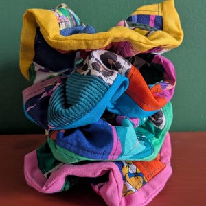 Può includere: Una pila di colorati elastici per capelli. Ogni elastico presenta un motivo e una combinazione di colori unici, tra cui giallo, blu, rosa, verde e arancione. I tessuti presentano vari disegni, come floreali, animalier e geometrici.