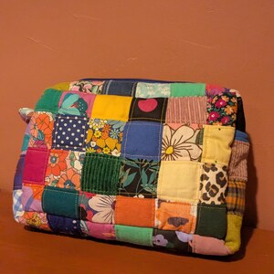 Può includere: Una pochette rettangolare con cerniera realizzata con un patchwork di vari quadrati di tessuto. Il design presenta un mix di colori a tinta unita tra cui giallo, arancione, verde e blu, insieme a tessuti con motivi floreali, a pois e leopardati.