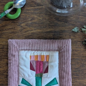 Op de afbeelding: Een handgemaakte gequilte onderzetter met een bloemenontwerp in roze, groen en oranje, op een houten oppervlak. Een glazen theepot met een metalen infuser en een groene lepelhouder zijn ook zichtbaar.