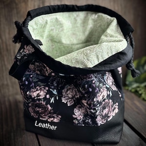 May include: Black drawstring bag with a floral peony print. The bag has a black leather base and a light green floral patterned interior. The word "Leather" is printed on the front of the bag.