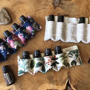 May include: Various essential oil holders with bottles are arranged on a wooden surface. The holders are made of fabric with floral and botanical patterns. One holder has a navy blue background with pink and white flowers. Another has a beige background with white leaf designs.